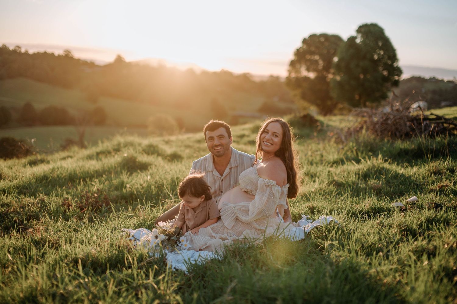 A family sits together on a blanket in a sunny meadow during golden hour while sun flares illuminate the scene.