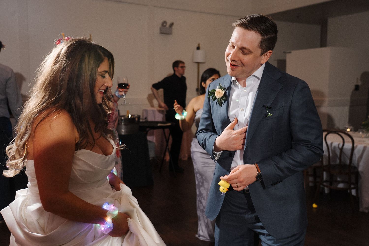A bride in a white gown dances energetically with a wedding guest in a navy suit at an indoor wedding reception.