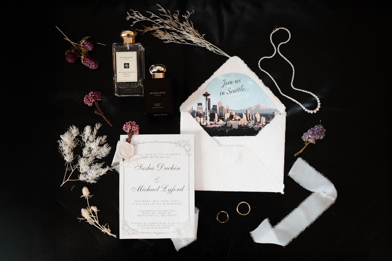 Elegant wedding rings photographed artistically with calligraphy invitations and dried flowers against a dark background.