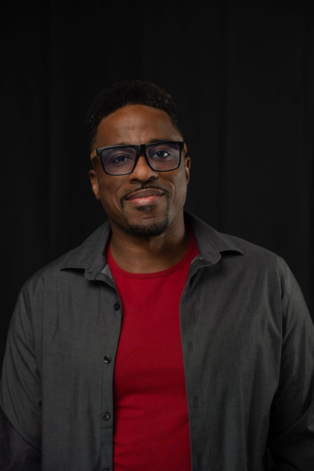 An African-american actor wearing a red shirt and gray jacket poses against a dark background in a professional portrait series.
