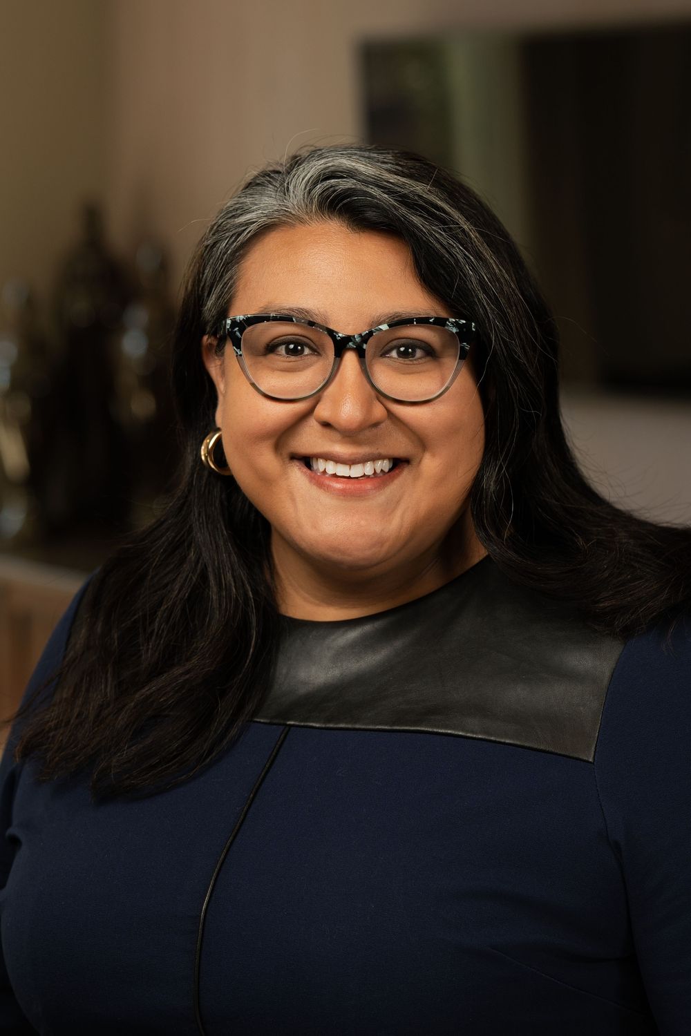 Professional headshot of a person with long dark hair wearing glasses and a navy and black color block top.