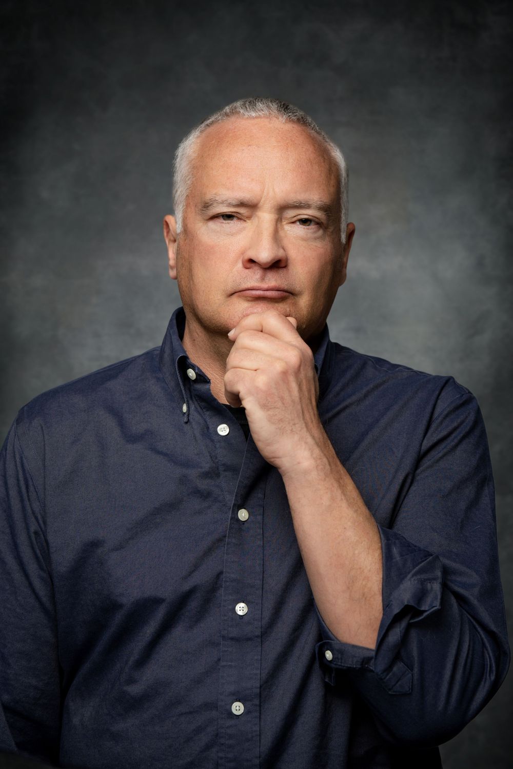 Professional studio portrait of a person in a navy button-down shirt with hand thoughtfully placed on chin against gray backdrop.