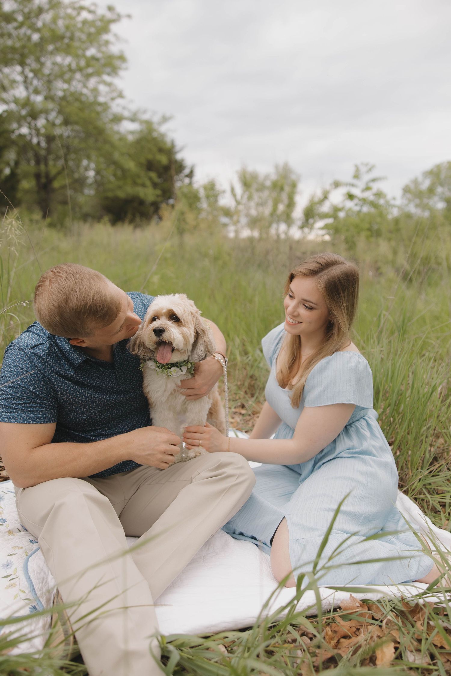 Couple relaxing on a white blanket in a grassy meadow with their small fluffy dog on a cloudy day.