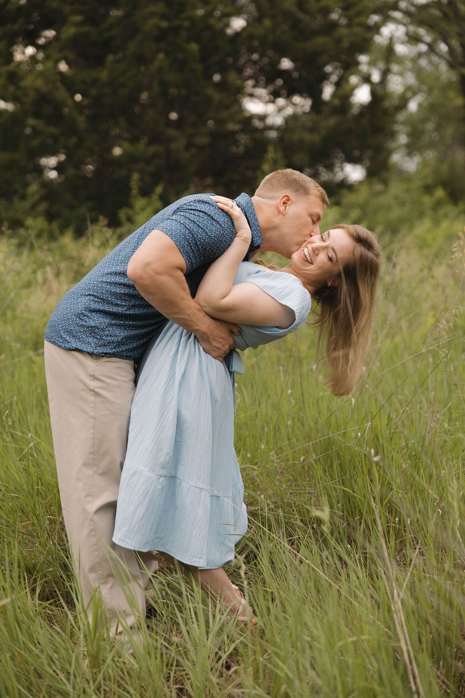 A romantic couple embraces in a sunlit meadow with tall grass, the woman wearing a light blue dress and man in blue shirt.