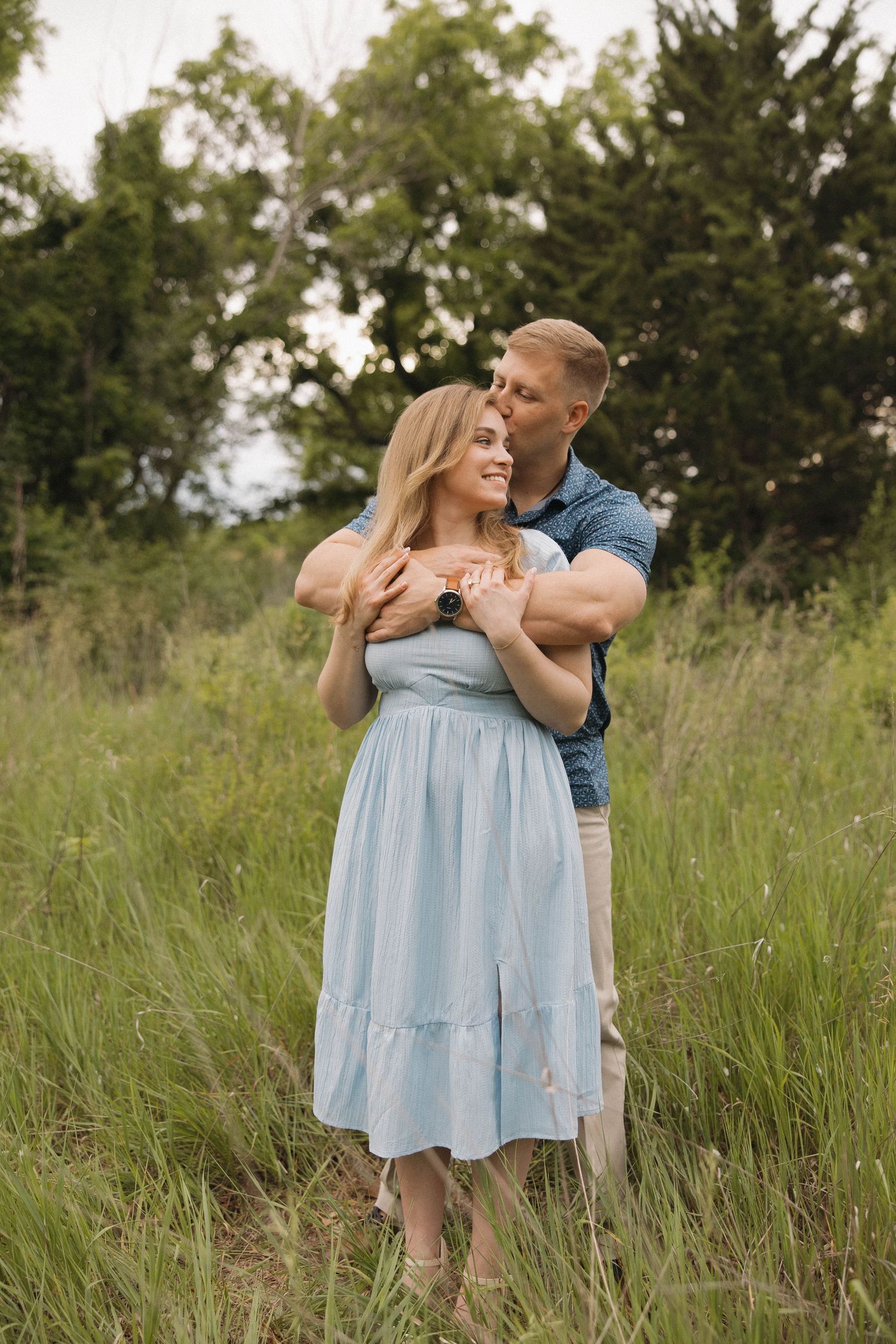 Romantic couple embraces in a sunlit meadow, with her wearing a light blue dress against a backdrop of green trees.