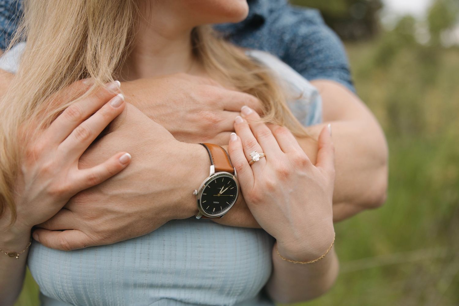 Close-up shot of embracing couple's hands with black wristwatch against light blue clothing in outdoor setting.