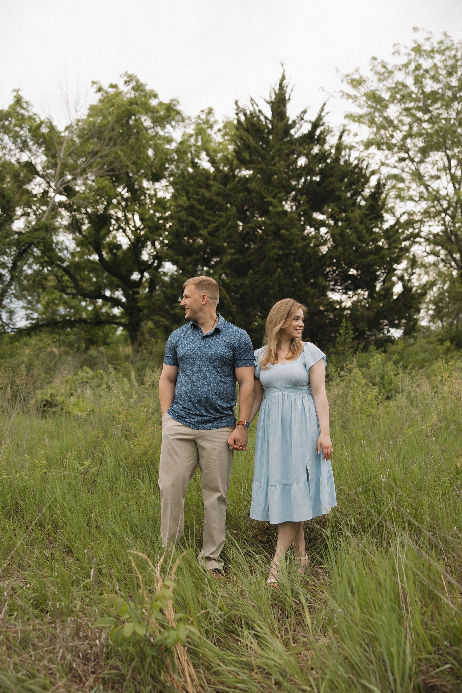 A couple stands together holding hands in a grassy field with trees in the background on a summer evening.