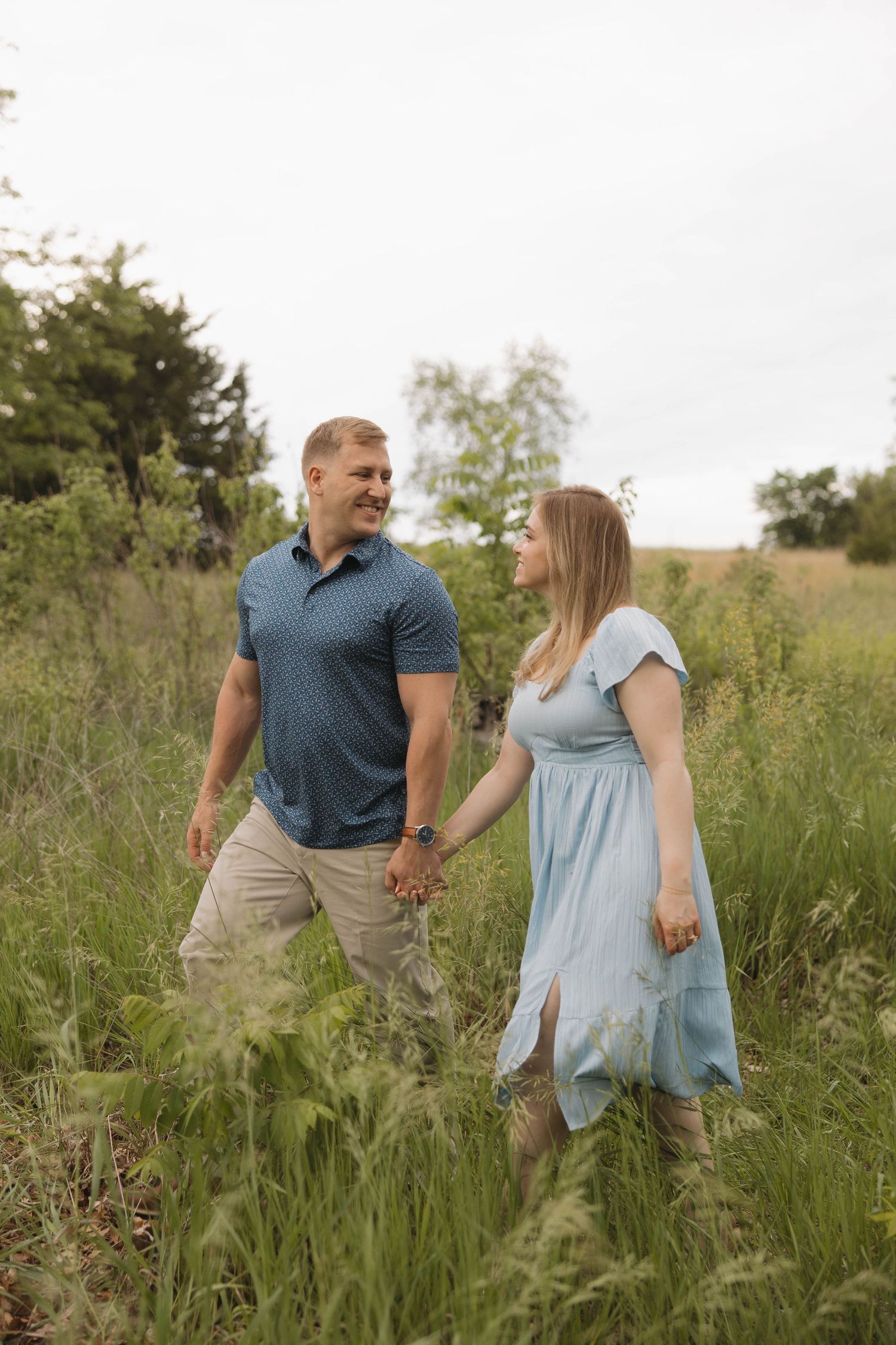 A couple holds hands while walking through a tall grass meadow on a warm summer day.