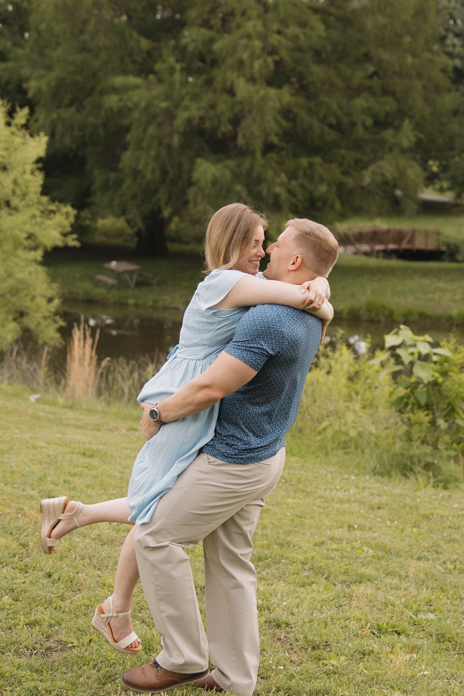 A romantic outdoor embrace in front of a tranquil pond, with sunlit trees creating a natural backdrop in the park.