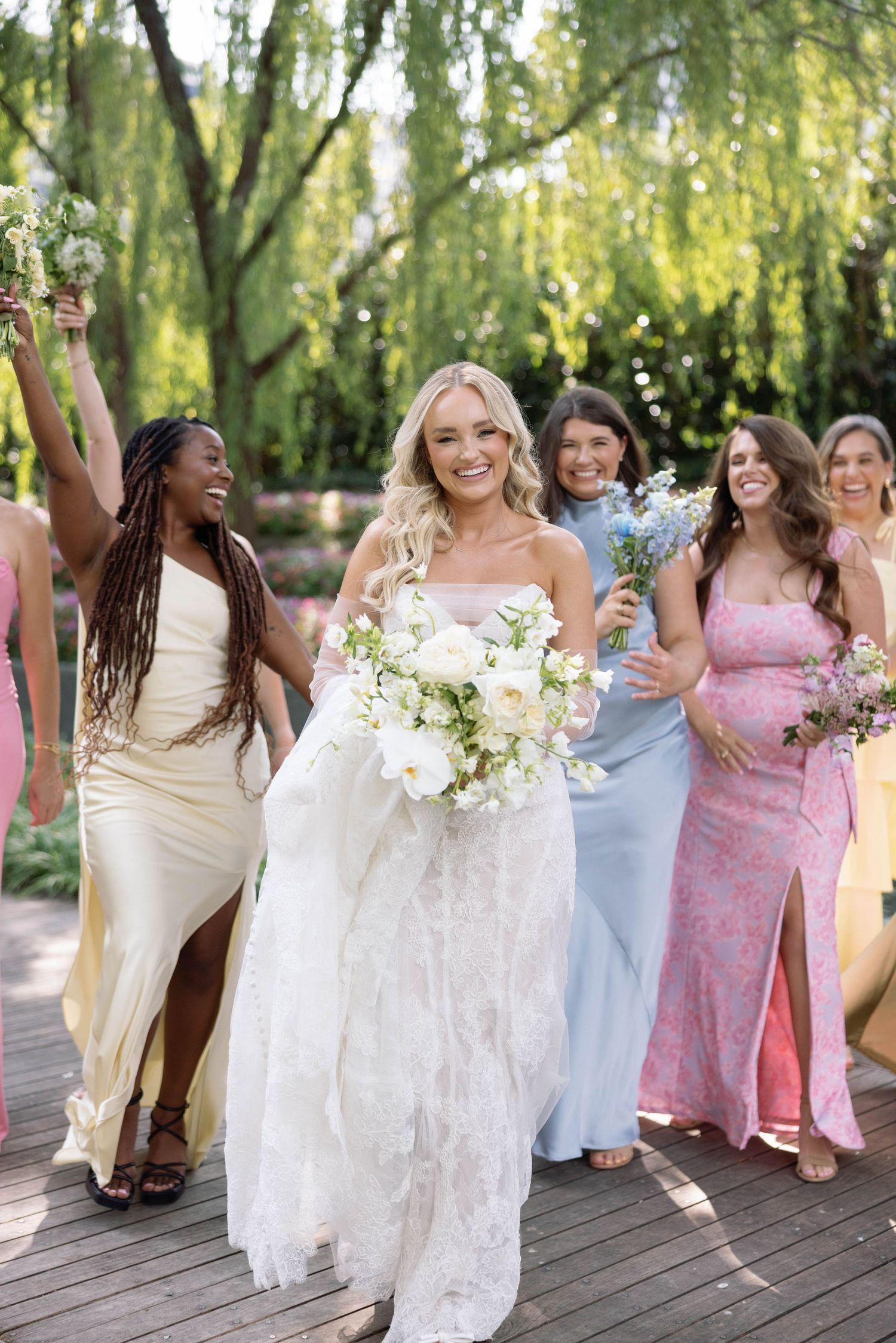 A bridal party in pastel and white dresses walks along a wooden deck under weeping willow trees.