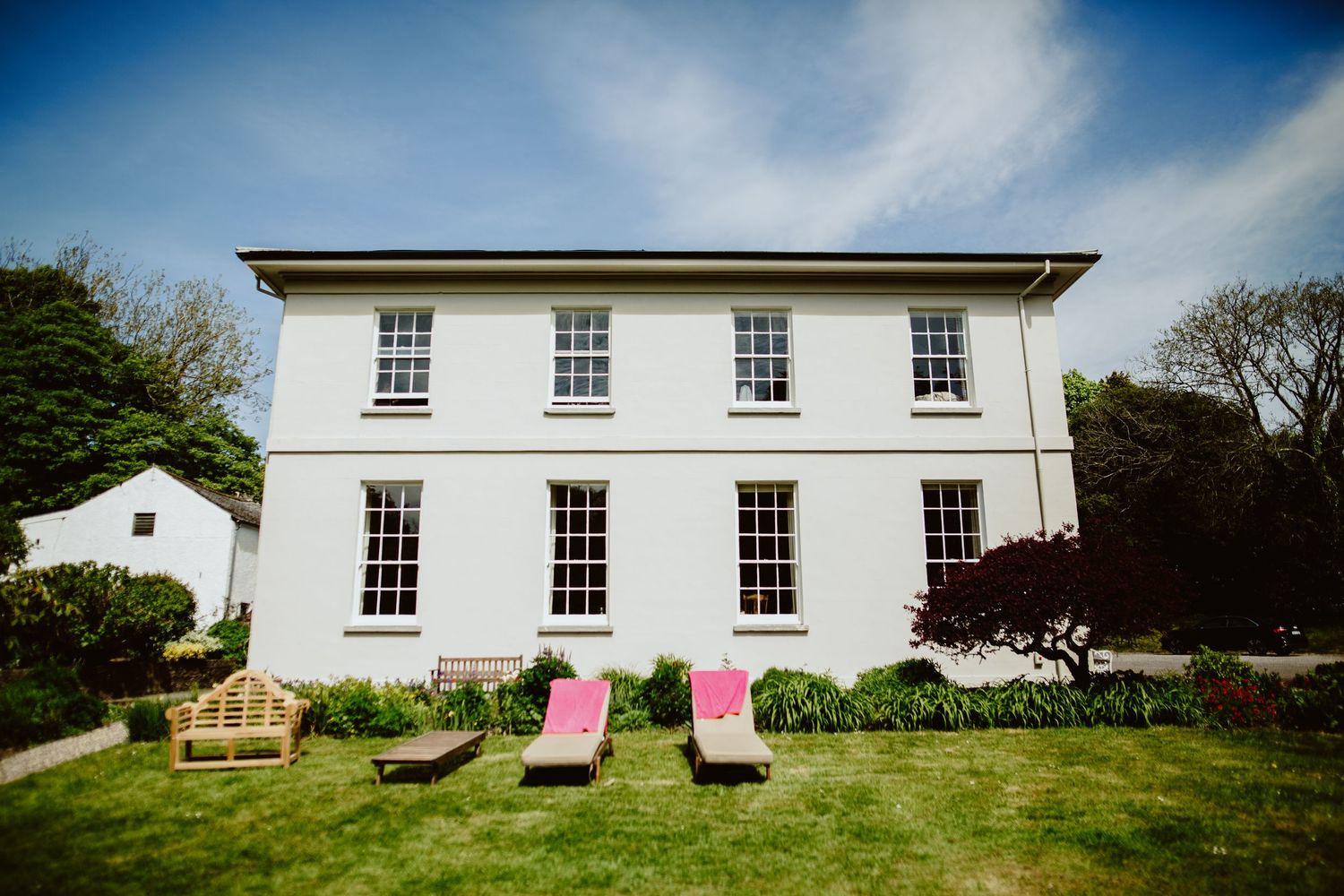A white Georgian style house with symmetrical windows and pink lounge chairs on a manicured lawn.