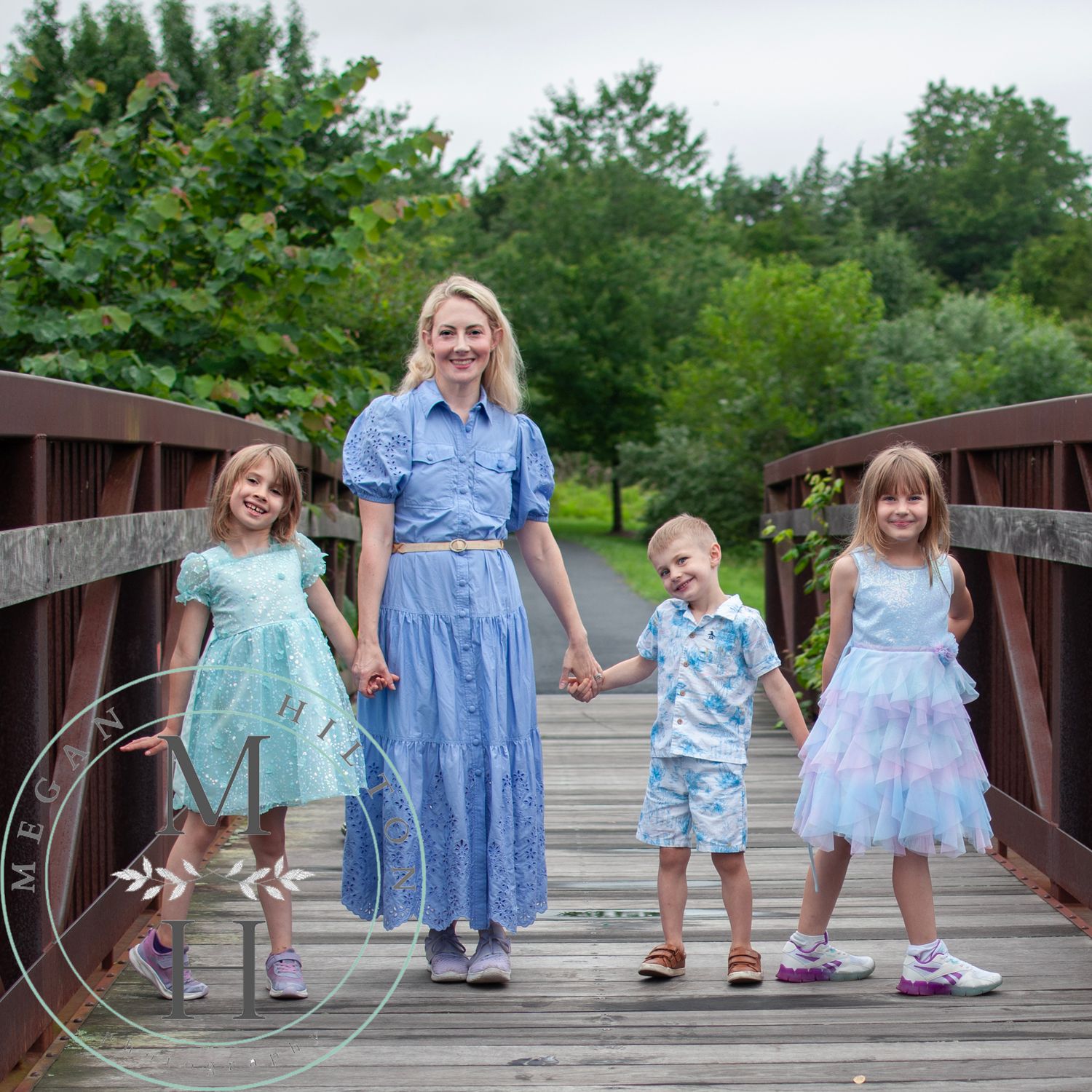 Family coordinated in blue and pastel outfits stands together on an iron bridge surrounded by greenery at the Broadlands Wetlands Preserve.