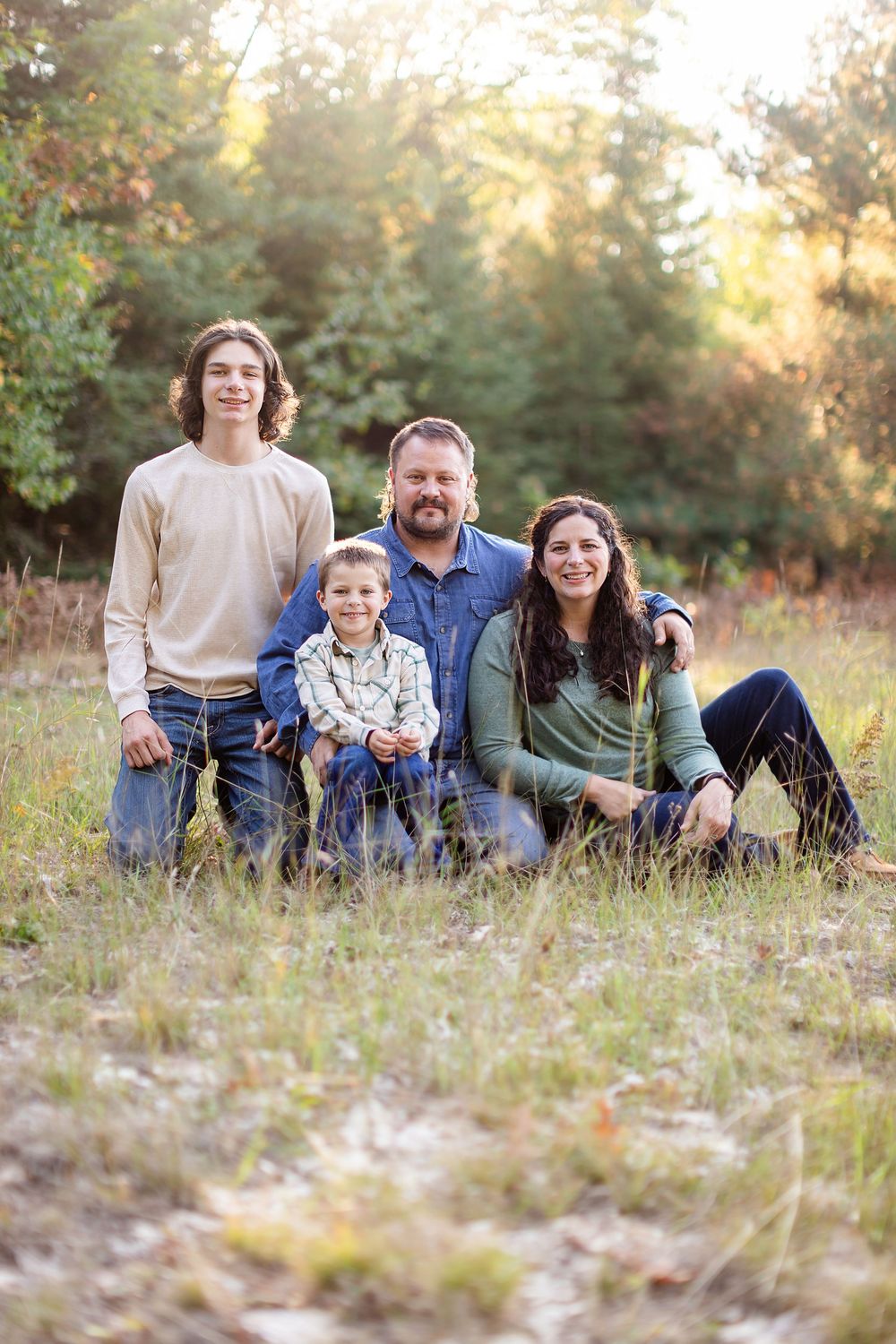 Family relaxing together in a sunny meadow with golden light filtering through trees in the background.