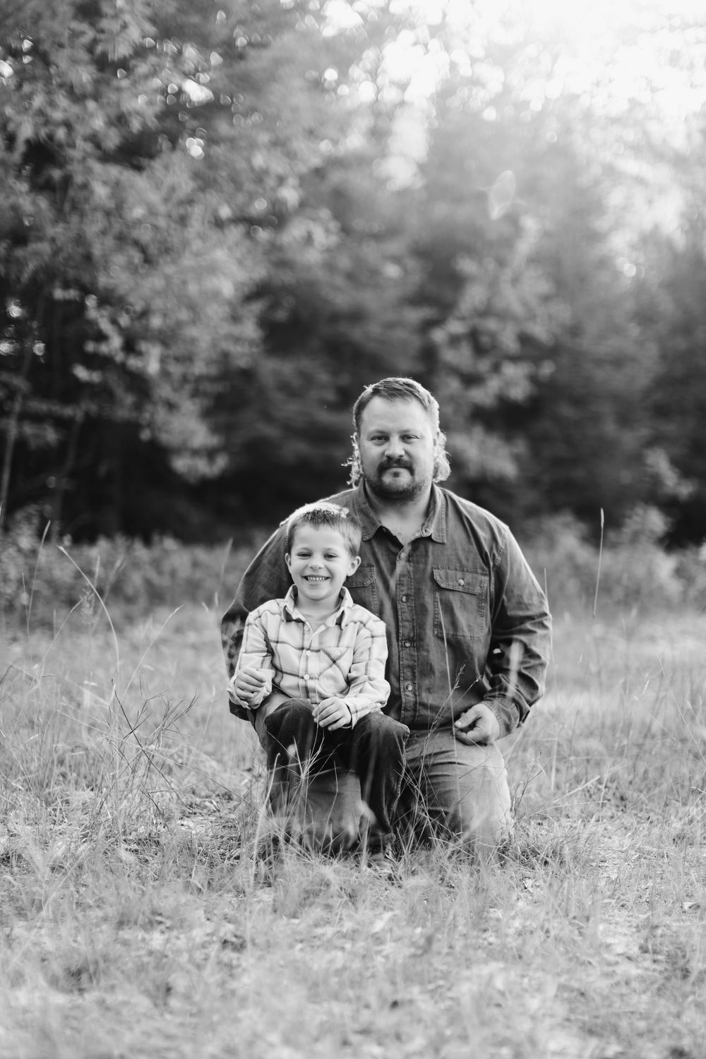 A black and white portrait of an adult and small child posing together in a grassy field with trees in background.