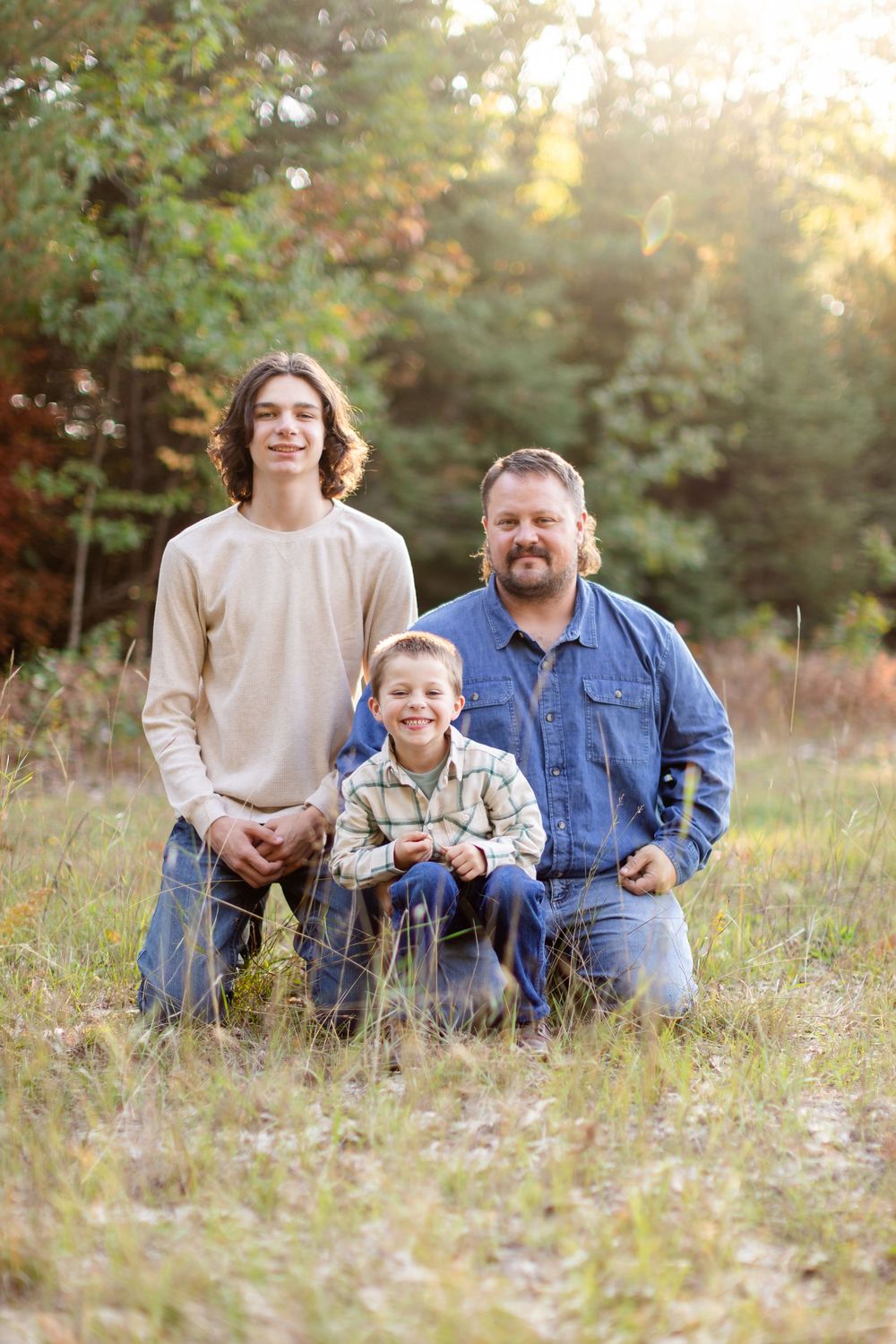A family portrait taken outdoors in a sun-drenched field with autumn foliage in the background.