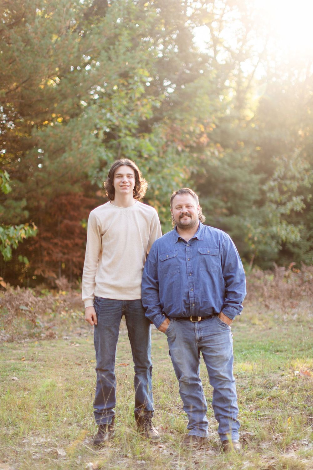 Two people stand side by side in casual attire during golden hour with autumn trees behind them.