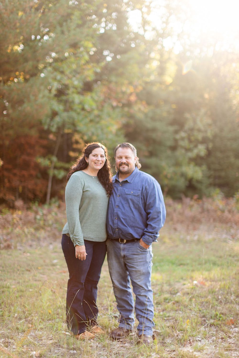 A couple poses together outdoors during sunset with fall foliage in the background.