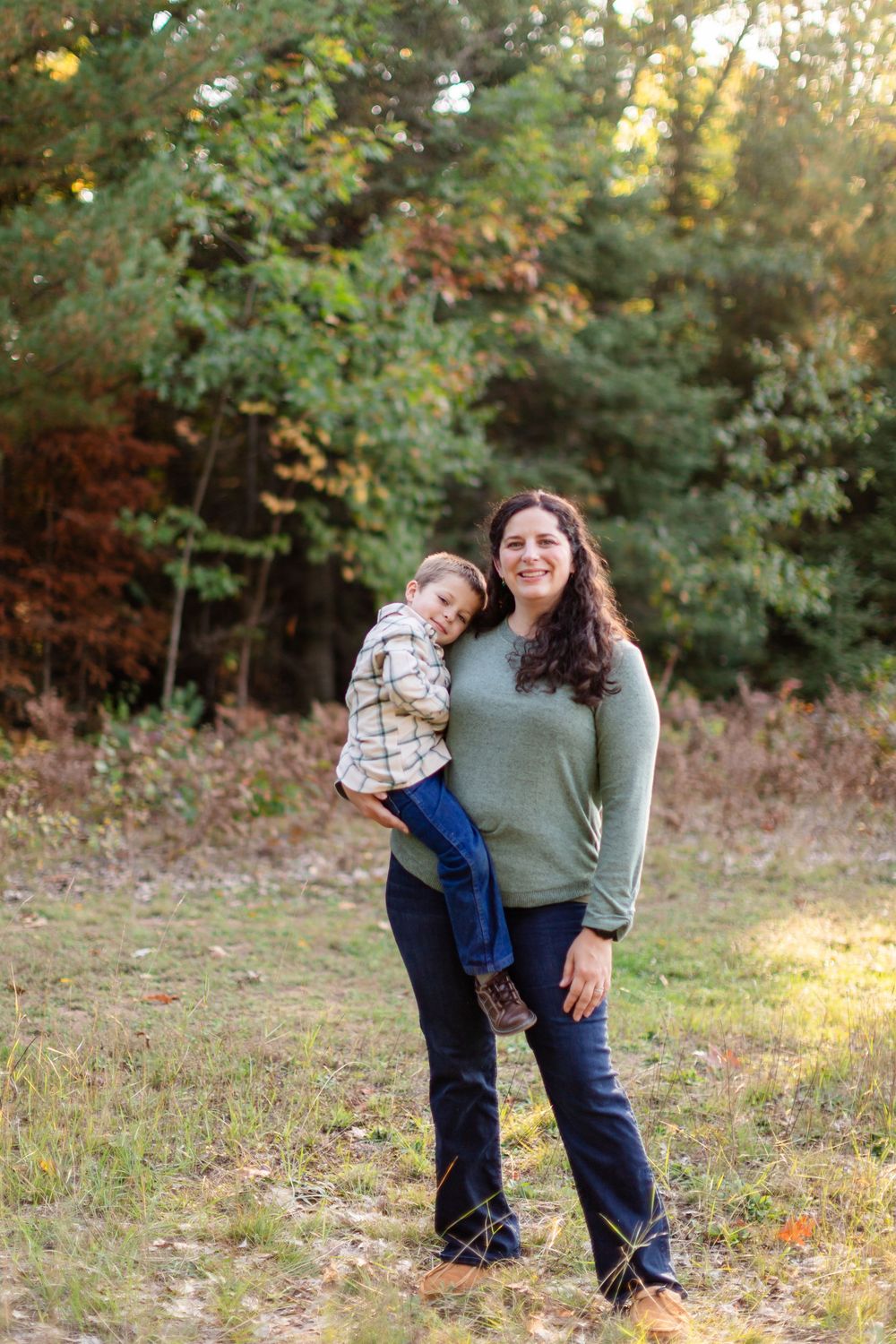 A parent and child share a playful moment while posing for a portrait in a wooded outdoor setting.