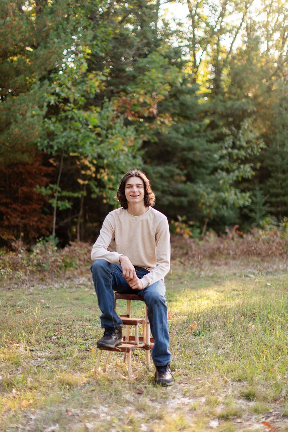 Person sitting on wooden chair in grassy field with autumn trees in background during sunset.