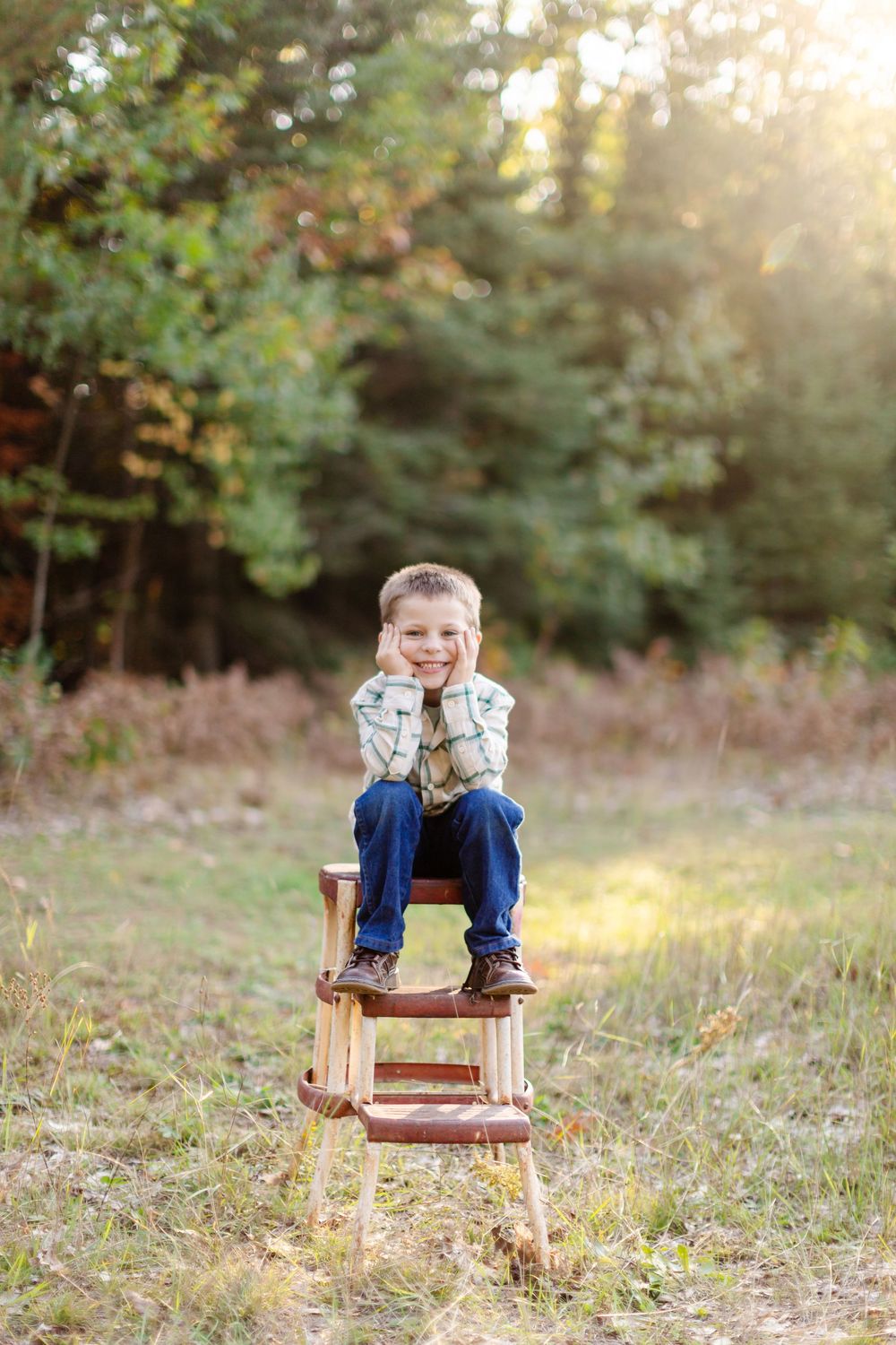 A young child sits thoughtfully on a wooden stool in an autumn field with sunlit trees in background.