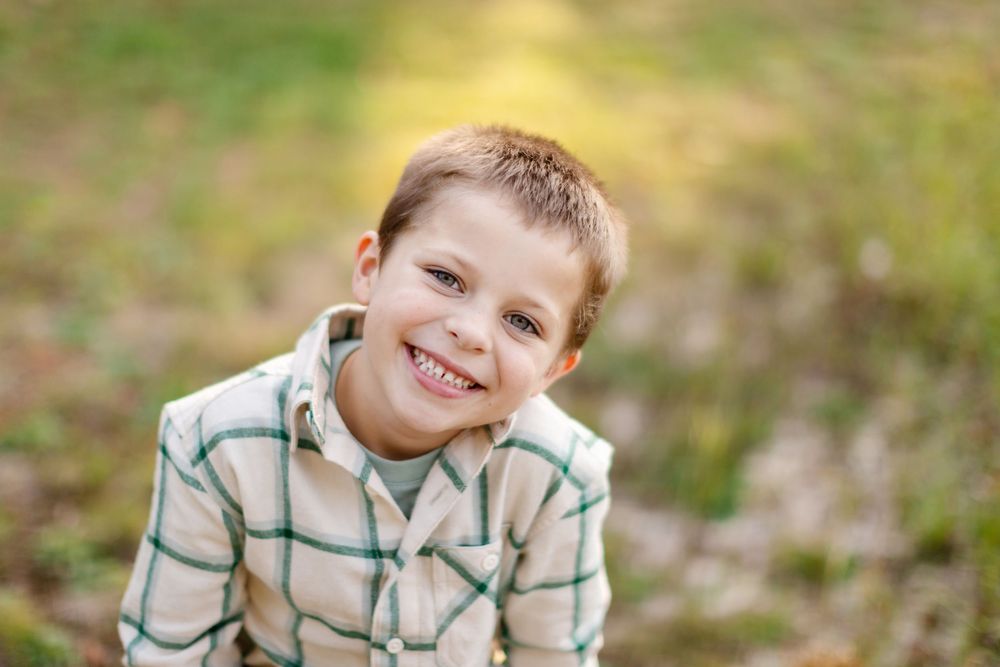 A child wearing a plaid shirt smiles warmly at the camera during an outdoor photo session.