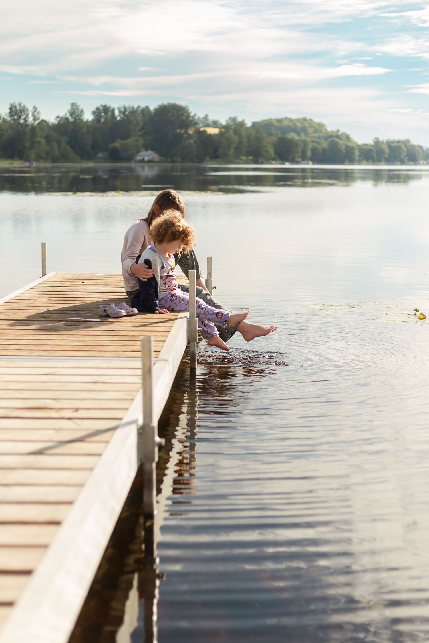 Two people sit with their feet dangling over a wooden dock on a serene lake during a peaceful summer day.