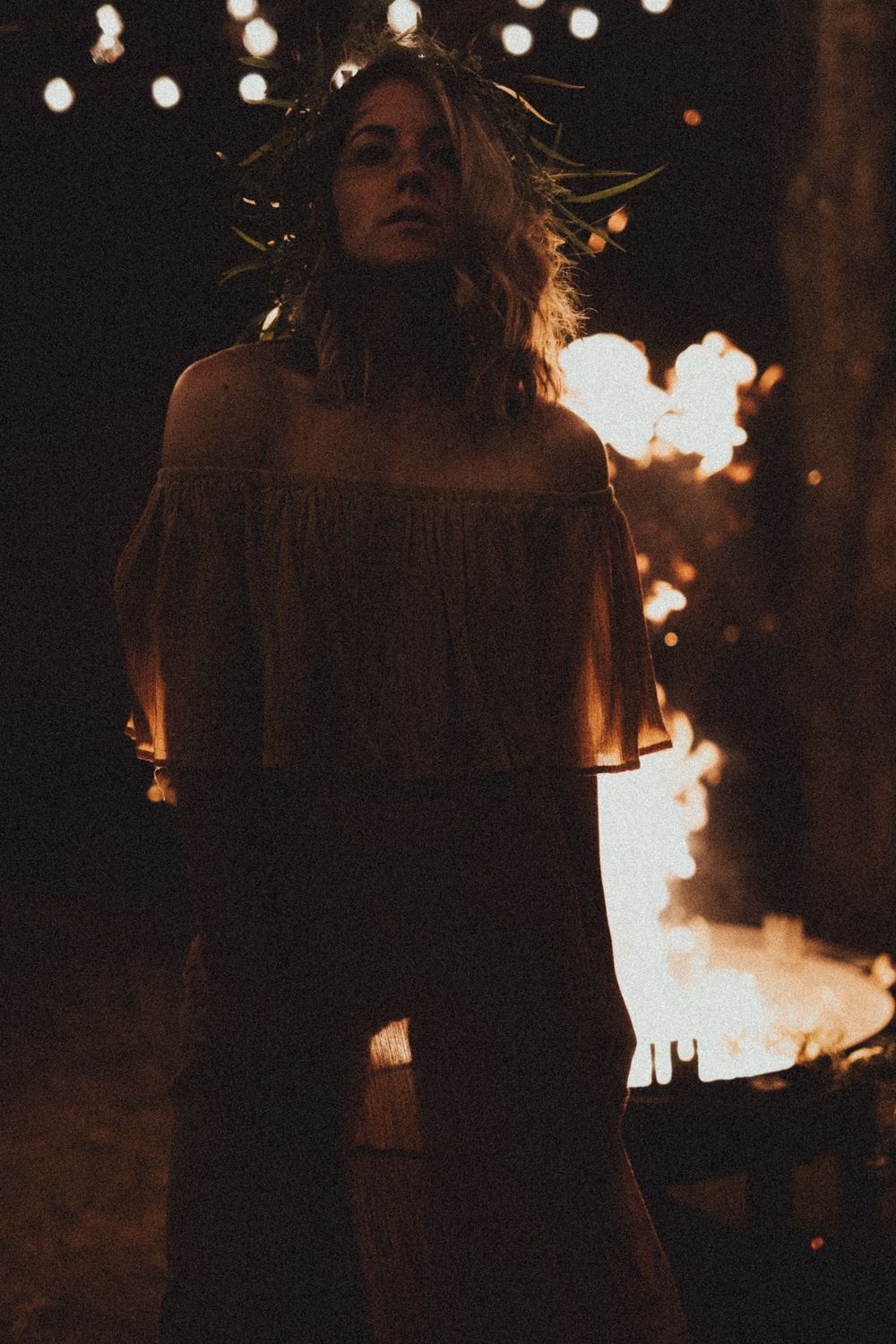 A figure stands in a dark moody setting with string lights overhead and warm firelight glowing in the background.