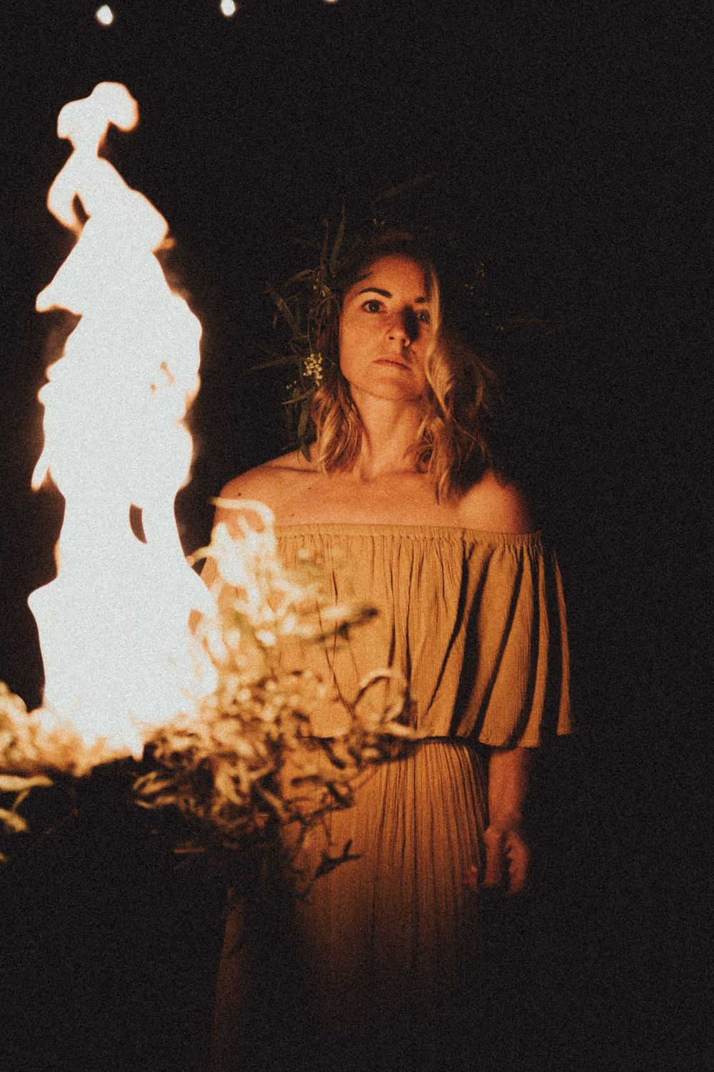 Dramatic portrait illuminated by firelight showing a figure in an off-shoulder dress standing beside glowing flames at night.