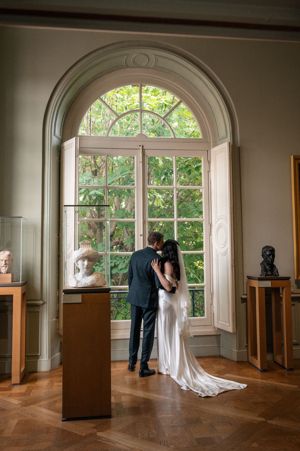 Wedding couple standing by large arched windows in a historic room with wooden floors.