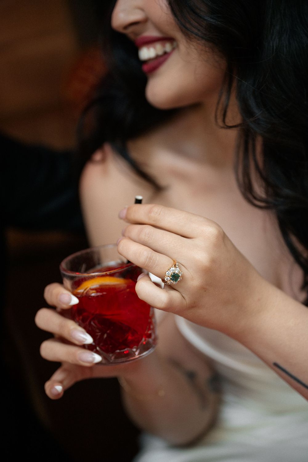 Close up of hands holding a red cocktail drink with manicured nails and ornate ring visible.