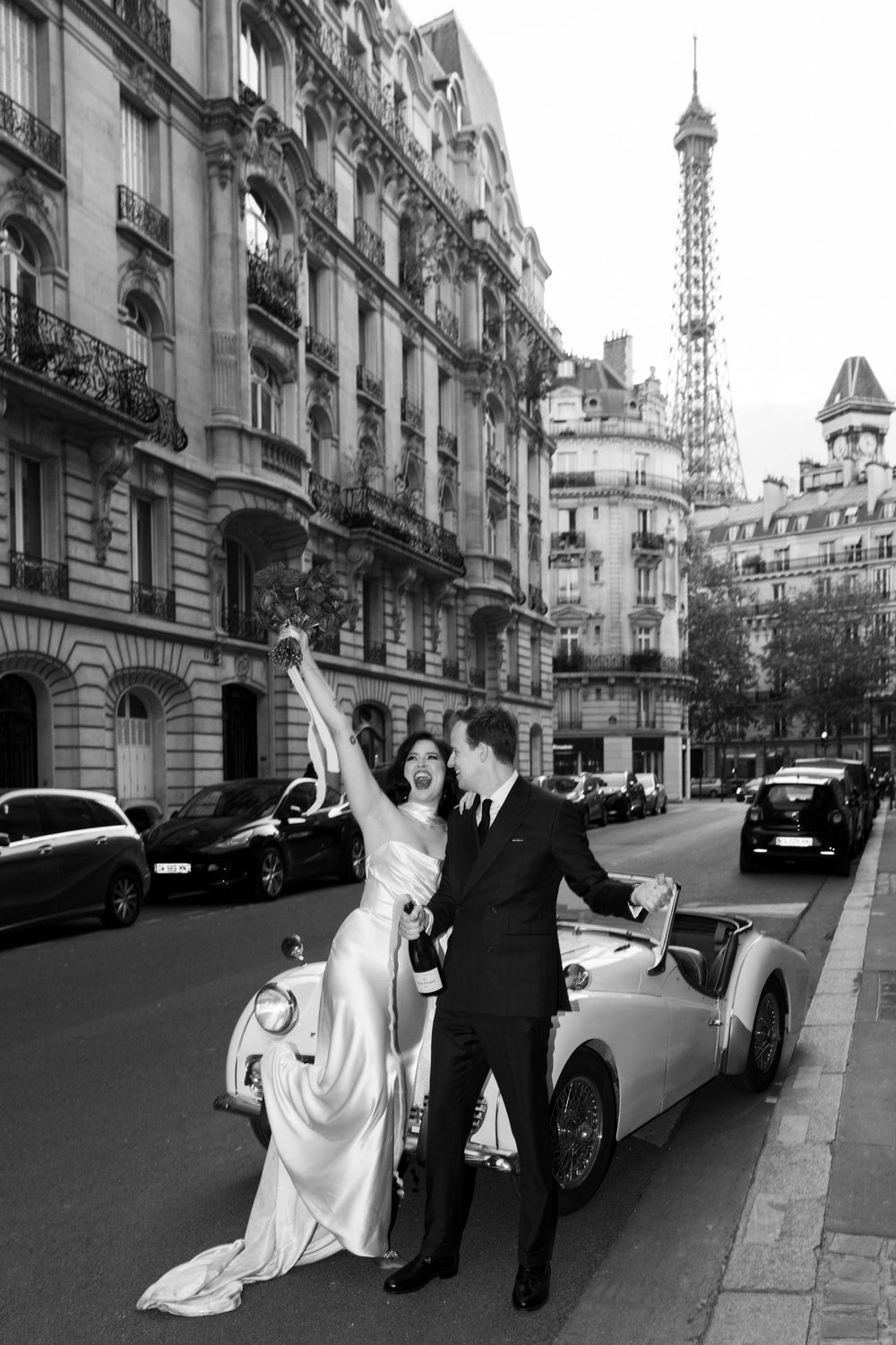 Vintage wedding scene in Paris with classic car and Eiffel Tower backdrop on elegant city street in black and white.