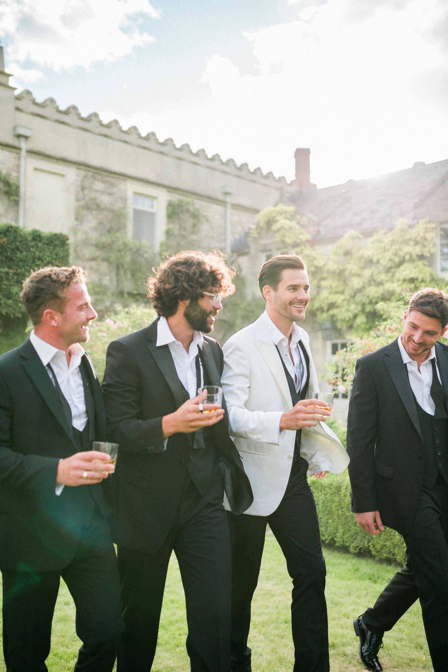 A group of groomsmen in black suits and a groom in white share laughs while holding drinks at an outdoor wedding. Euridge Manor Photography