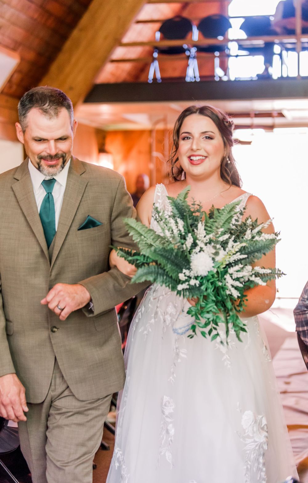A wedding couple walking together indoors, with the bride holding a lush green and white fern bouquet.