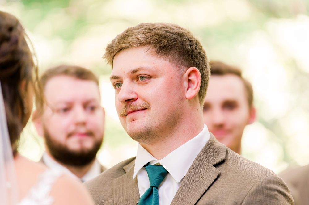 A person in a grey suit with a teal tie smiles warmly during what appears to be a wedding ceremony.
