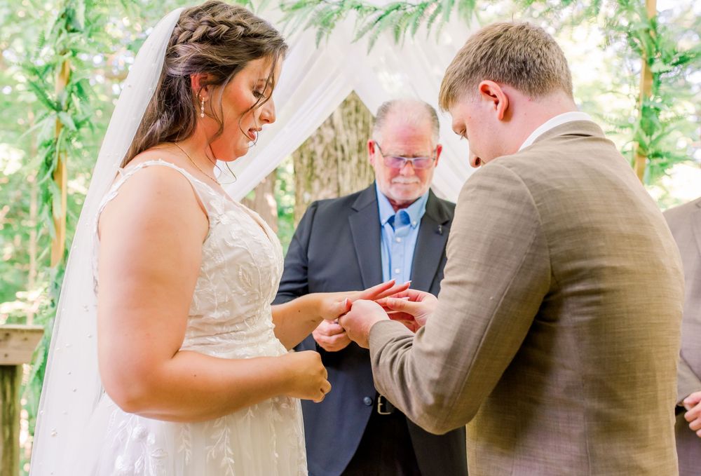 A couple exchanges rings during an outdoor wedding ceremony beneath a leafy arbor with a minister presiding.