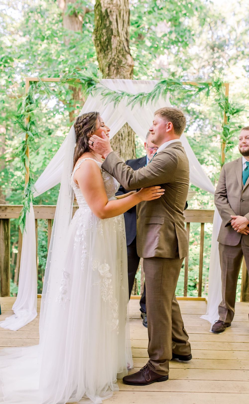 A romantic wedding ceremony takes place on a wooden deck adorned with greenery and white tulle decorations.