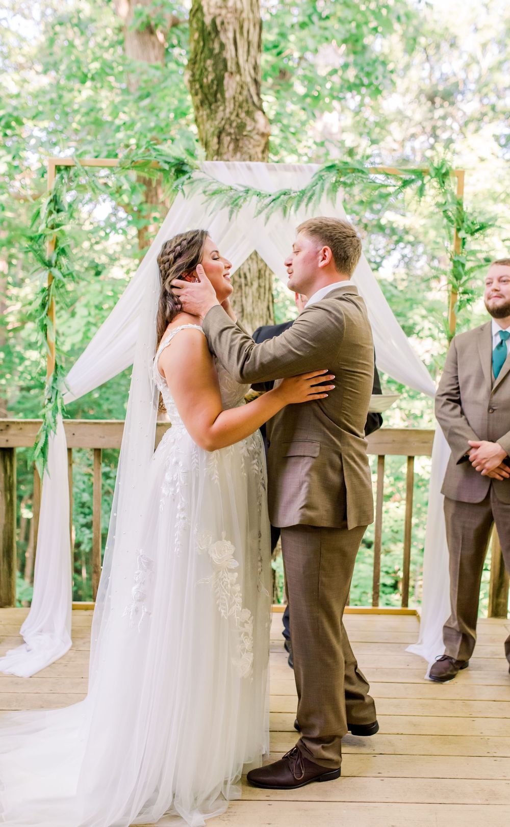 Happy couple sharing a playful first kiss moment during their outdoor wedding ceremony on a wooden deck surrounded by greenery.
