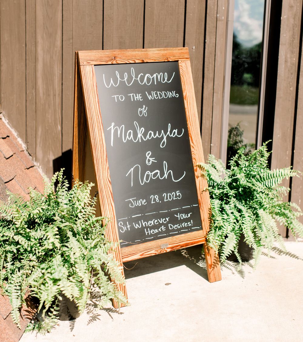 A wooden framed chalkboard wedding welcome sign with greenery displayed against a rustic wood wall.