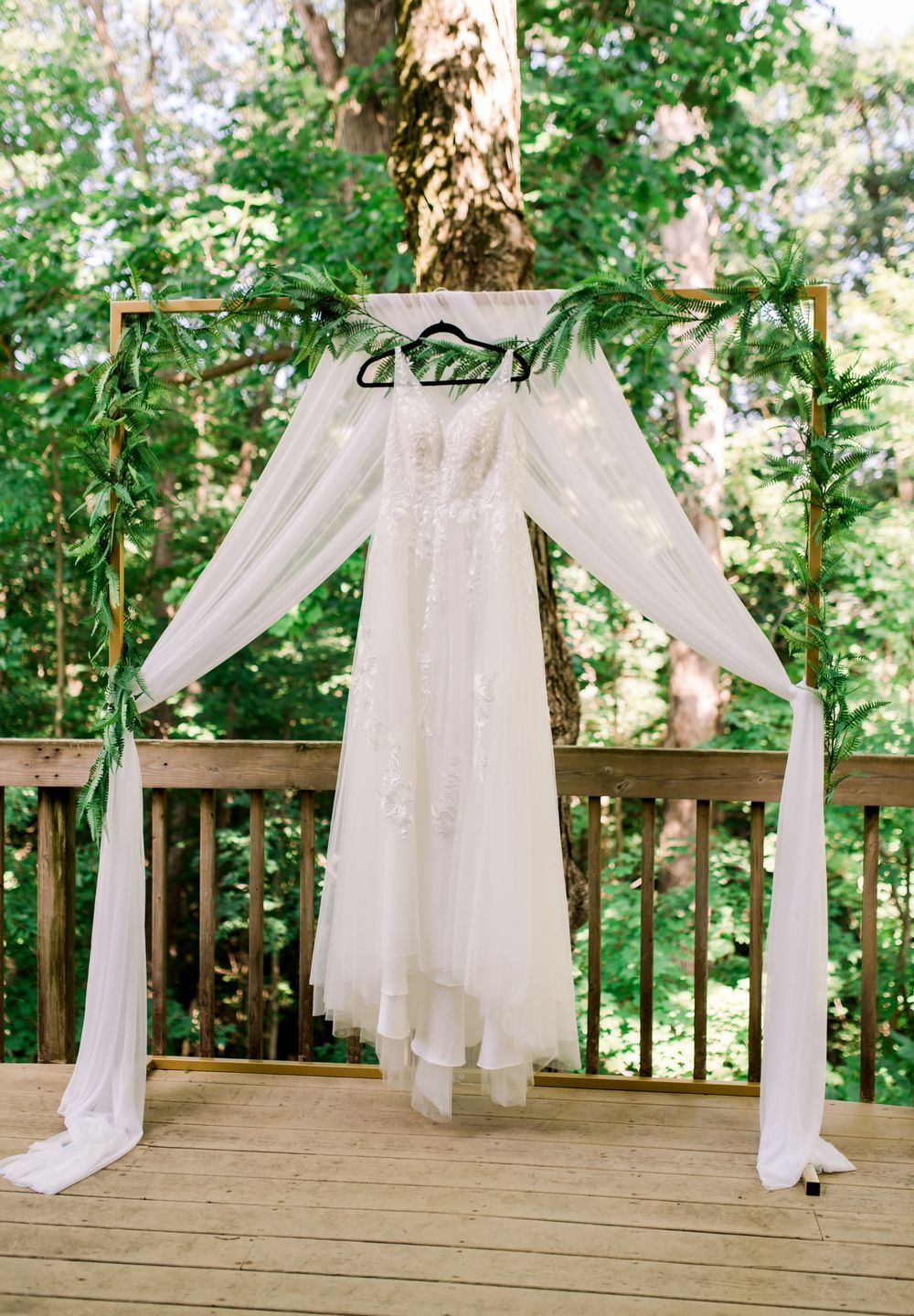 Elegant white wedding dress with long sleeves hanging from draped fabric canopy on an outdoor wooden deck surrounded by trees.