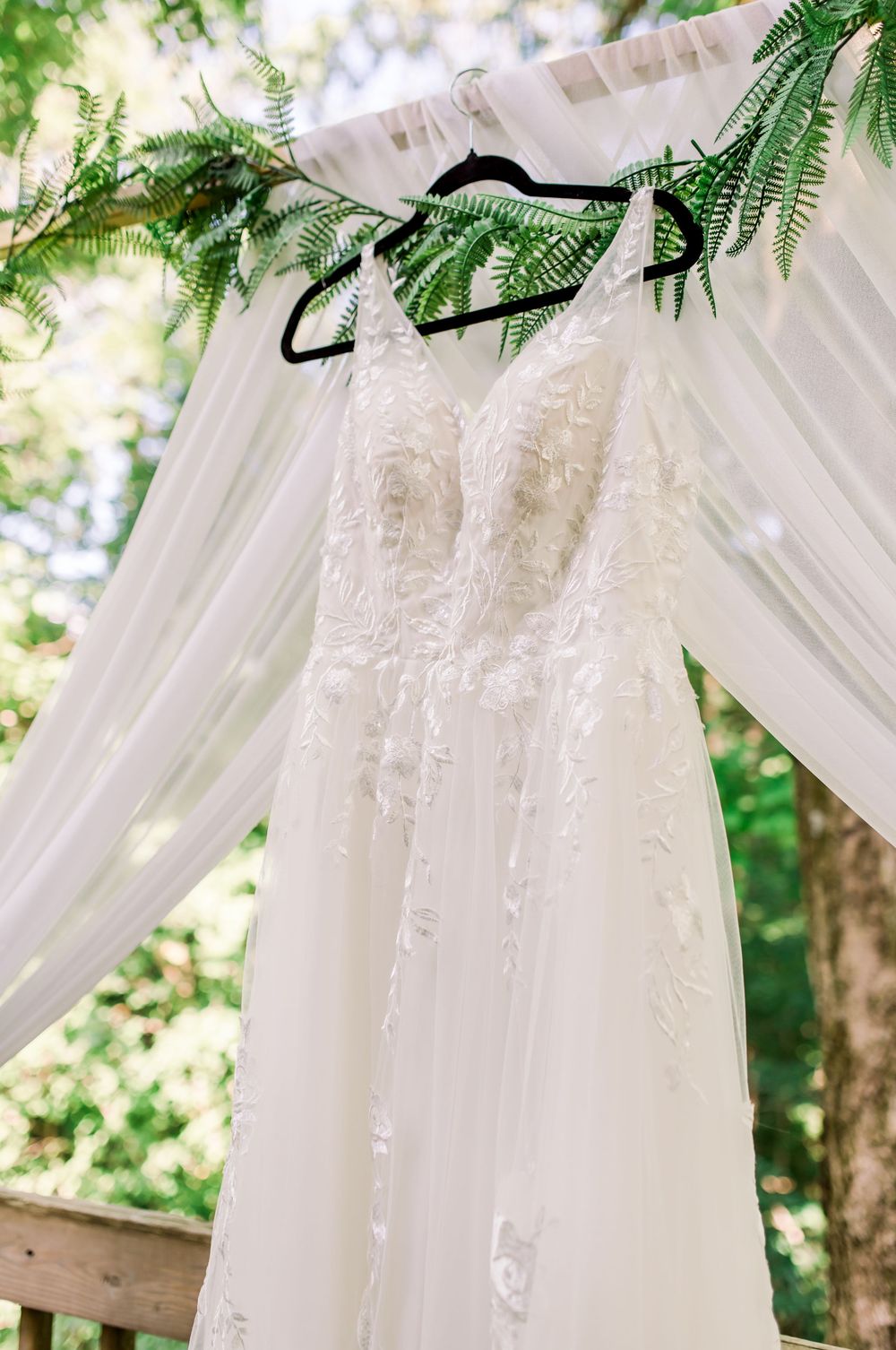 A delicate white lace wedding dress hangs from a black hanger against draped fabric and ferns in natural sunlight.