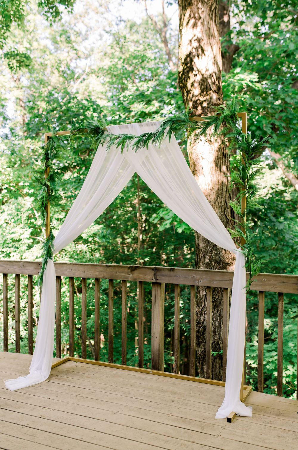 A wooden deck ceremony arch draped with white sheer fabric and greenery in a forest setting.
