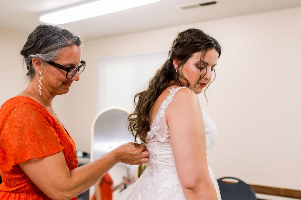 Someone in a bright orange top helps fasten the back of a white wedding dress during bridal preparations.