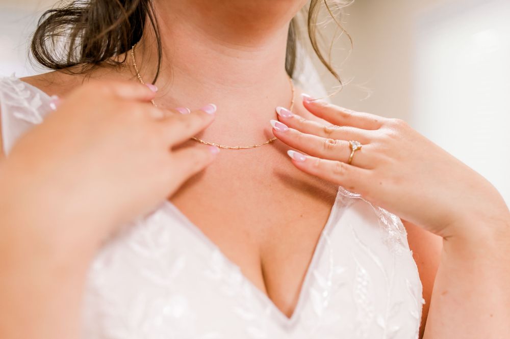 Close up of hands wearing delicate rings and necklace against white lace wedding dress bodice.