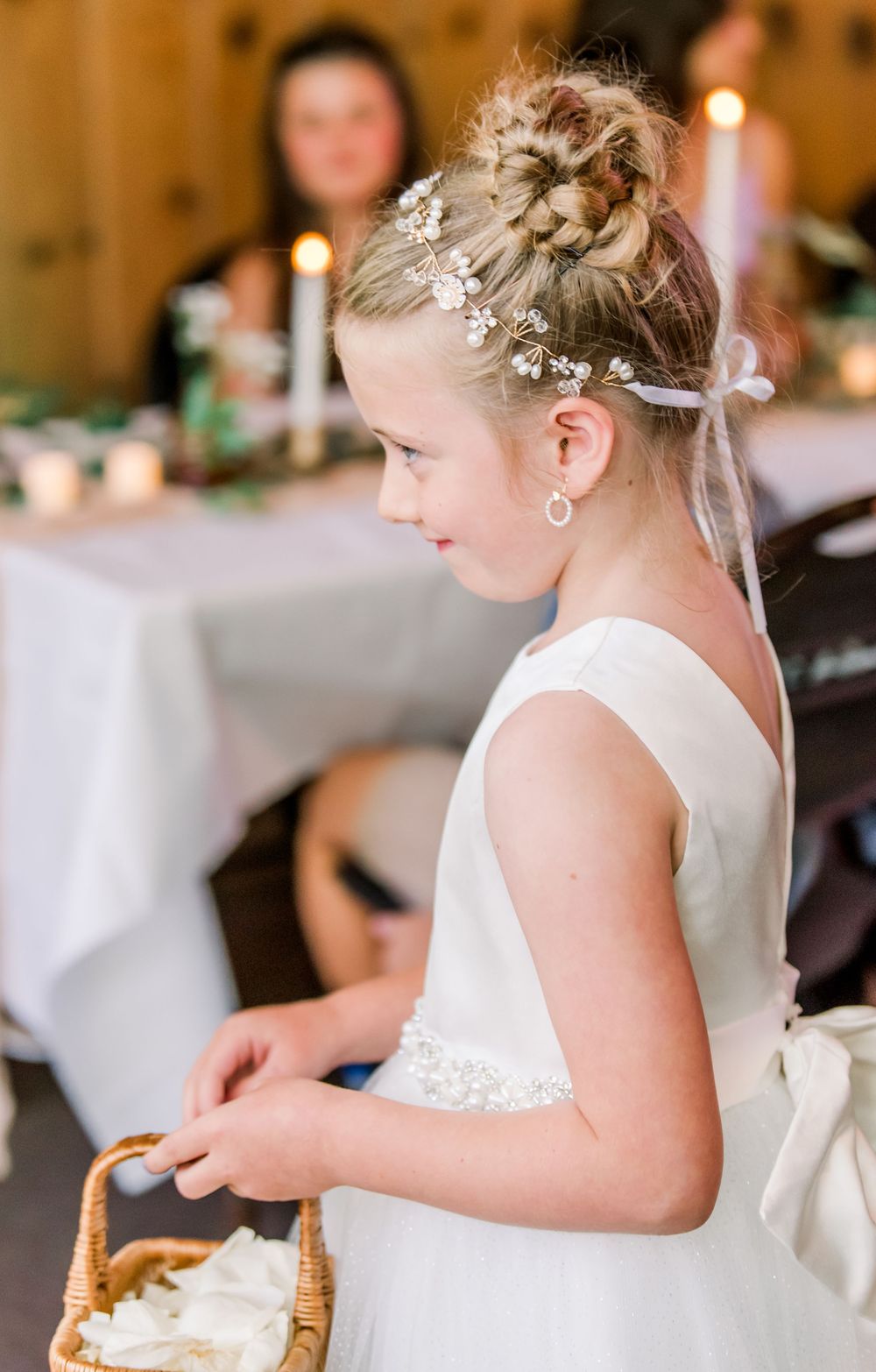 Young girl in white formal dress with braided updo and floral hair accessories at wedding reception.