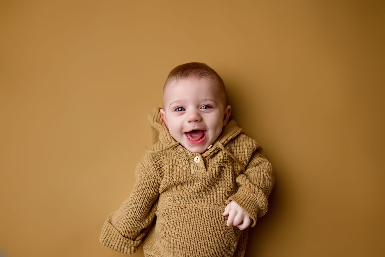 Laughing baby wearing a cozy ribbed brown sweater lies against brown background in studio portrait.