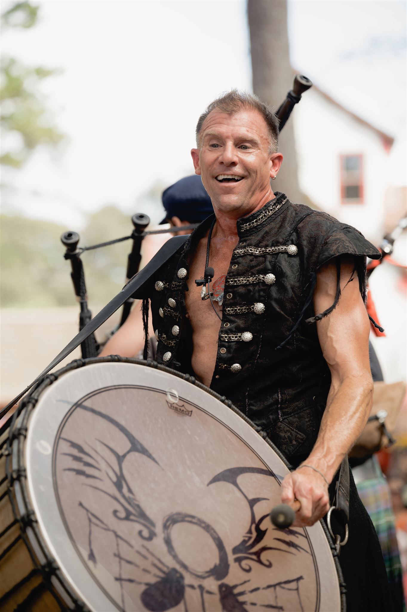A muscular performer in a black military-style vest plays a large decorated drum during an outdoor performance.