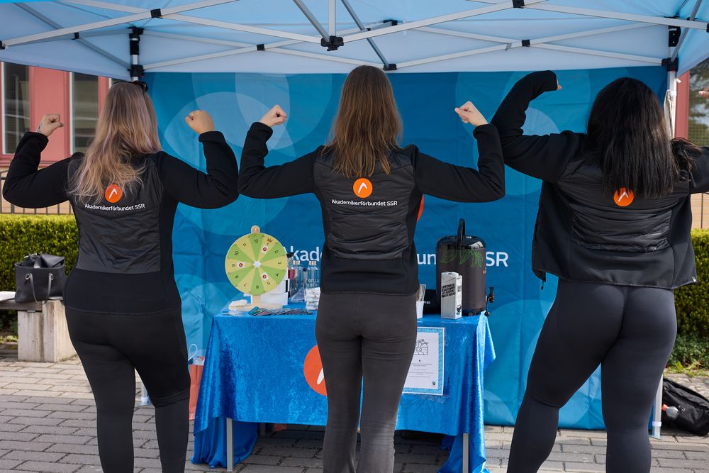 Three people in matching black jackets stand with arms raised at an outdoor event booth with blue backdrop.