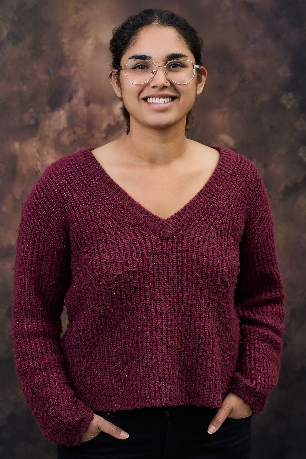 A warm portrait shows a person in a burgundy v-neck sweater smiling in front of a mottled brown backdrop.