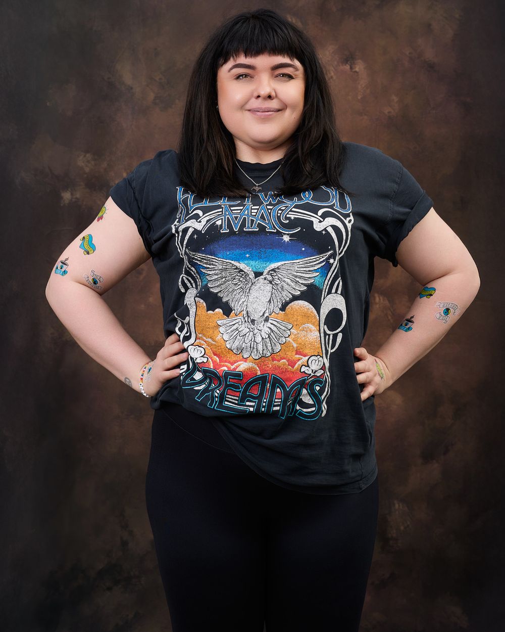 A person wearing a heavy metal t-shirt with an eagle design poses with hands on hips against a dark backdrop.