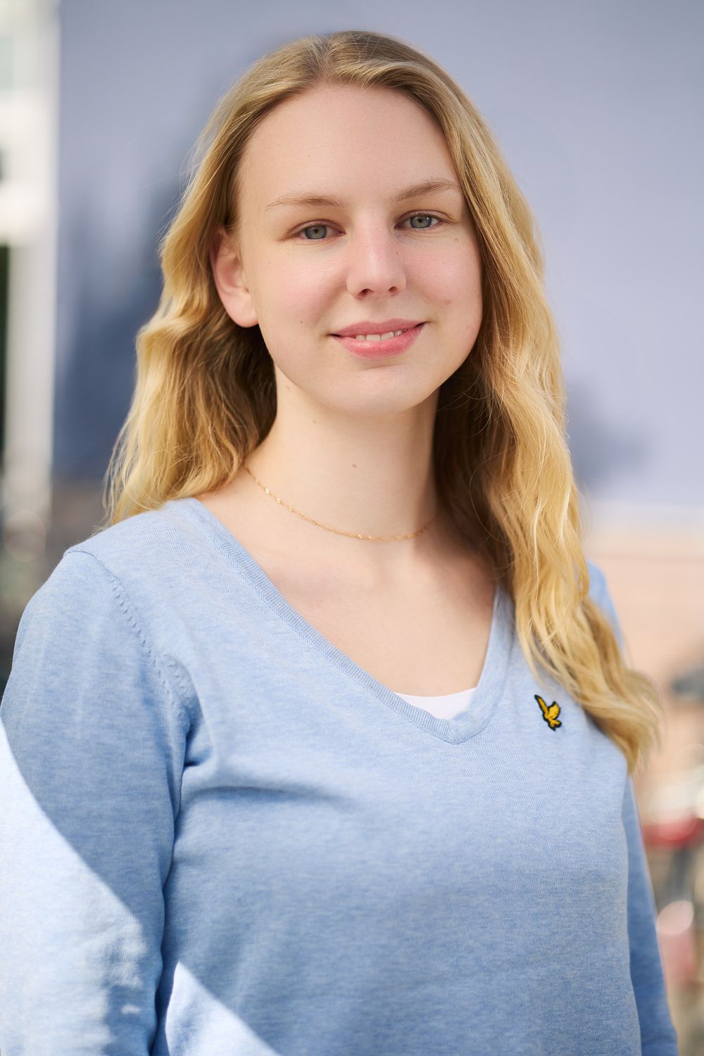 Young woman in light blue v-neck sweater with long blonde hair smiling at camera against blurred background.